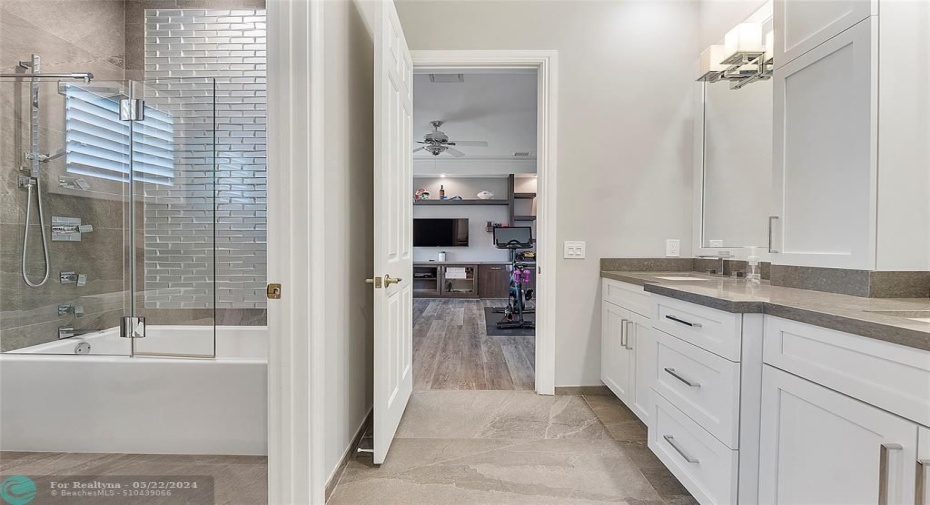 Jack & Jill modern bathroom with grey porcelain flooring and white vanities & accent wall and soaking tub, leading into the fitness room with wooden look porcelain flooring